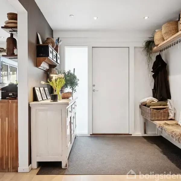 Dør står i midten af en entré med træmøbler og pæne opbevaringskurve. Planter og indrammede billeder dekorerer overfladerne, mens en bænk med puder er til højre.
