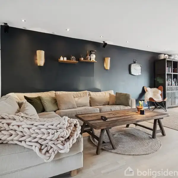 Stor beige sofa med strikket plaid, står mod en mørk væg. Træbord foran sofaen. Hylder med dekorative genstande hænger på væggen. Lyse, moderne stue med naturligt lysindfald.