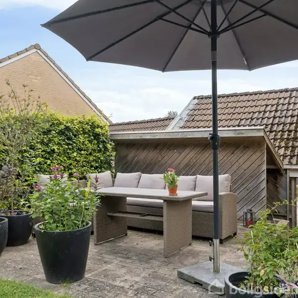Terrassemøbler står i en have, bestående af en sofa, et bord og en parasol. De er omringet af store potteplanter og en hæk, ved siden af en træbeklædt skurvæg.