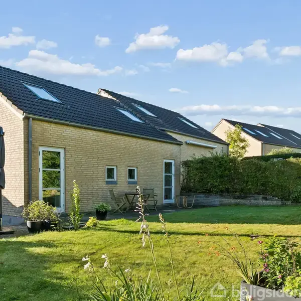 Et moderne, gult murstenshus med skråt, sort tag står i en velholdt have med græsplæne og blomsterbede. Ved huset ses havemøbler og en parasol. Blå himmel og nabohuse i baggrunden.