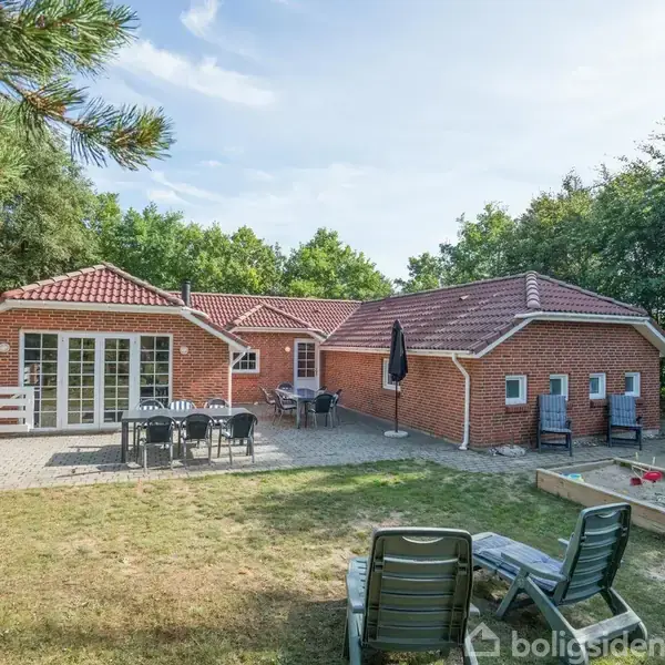 Et rødt murstenshus med rødt tegltag har en terrasse med havemøbler. Der er liggestole og en sandkasse i forhaven, omgivet af træer og græsplæne.