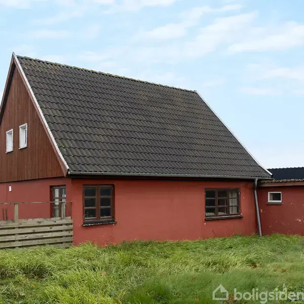 Et rødt hus med mørkt tag står stille i en græsbevokset have. Der er en lav træhegn foran og en klar blå himmel i baggrunden.
