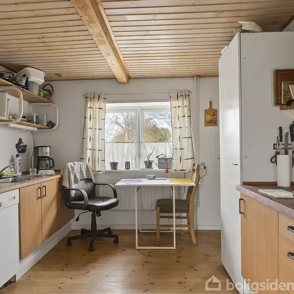 En lille køkken med træloft og gulv, rummer en køkkenbord med opvaskemaskine, hylder og køleskab. Et skrivebord og stol står ved et vindue med udsigt til naturen.