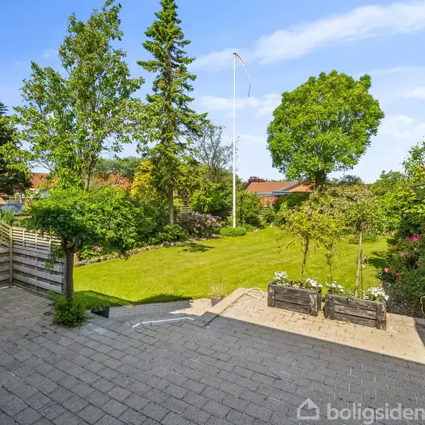 En flisebelagt terrasse med blomsterkasser. Handler om husets have i baggrunden, omgivet af træer og en flagstang. Solrigt vejr forstærker den grønne vegetation.