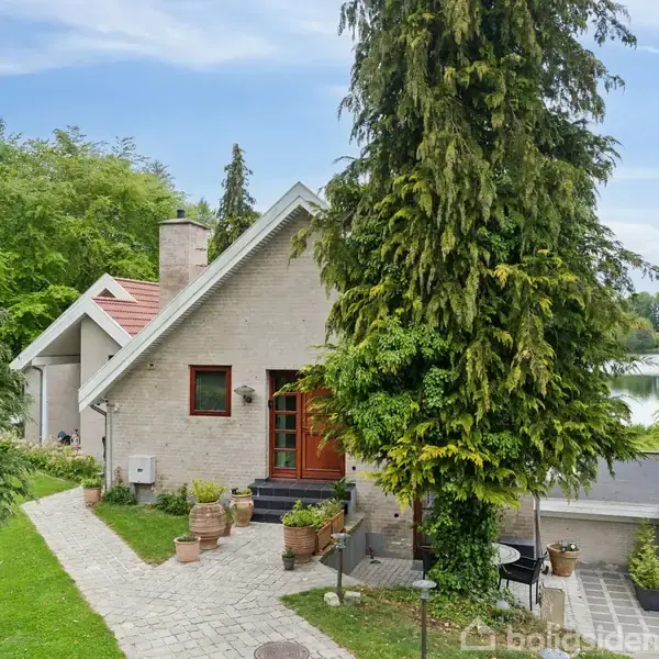 Et hvidt murstenshus med rødt tag ligger omkranset af træer og græsplæne. Foran huset fører en stensti til hoveddøren, omgivet af krukker og grønne planter.