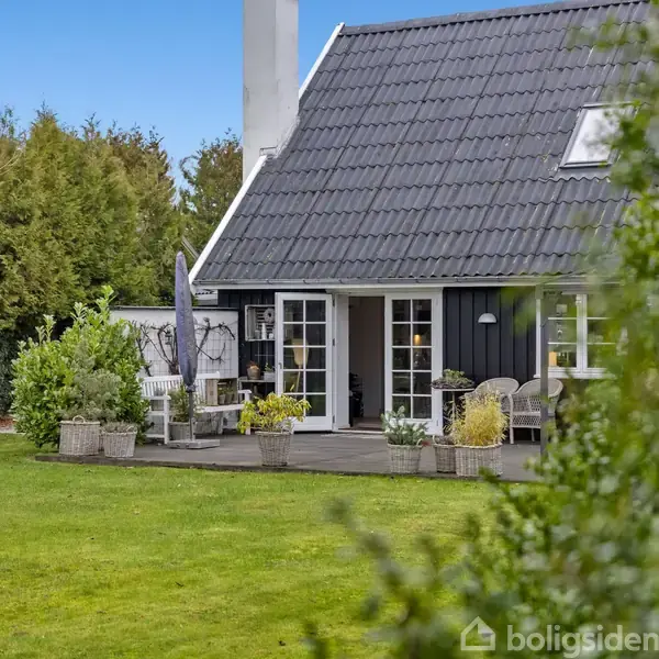 Hus med sort træfacade og hvidt tag står i forgrunden. Terrassen har havemøbler og planter. Omgivet af en græsplæne og træer på en klar dag.