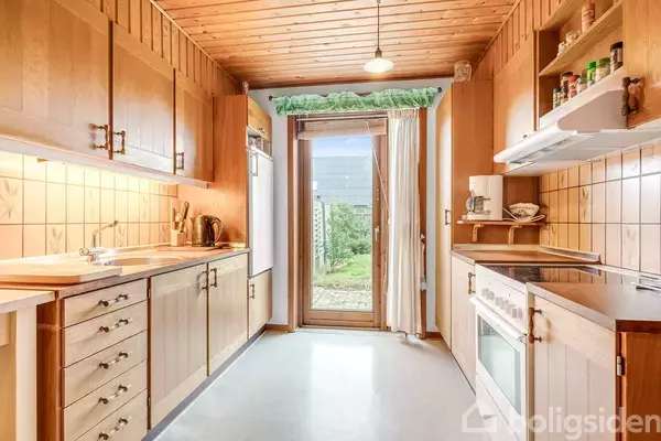 Et langt køkken med træskabe og skuffer. Rummet har hvidevarer, hylder med krydderier, og et stort vindue samt glasdør, der leder ud til en grøn have.