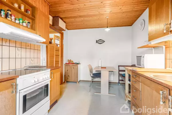 Køkken med en hvid komfur, træskabe og hylder med krydderi-dåser. Spisebord med stole står midt i rummet. Hvid mikrobølgeovn står på en træbordplade. Vægdekoration i form af en fisk.