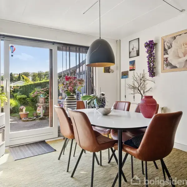 Et spisebord med seks brune stole står i en spisestue. Bordet er dekoreret med en vase og hænger under en sort lampe. Rummet har adgang til en terrasse med grønne planter. Veggen har billeder og en stor roseillustration.