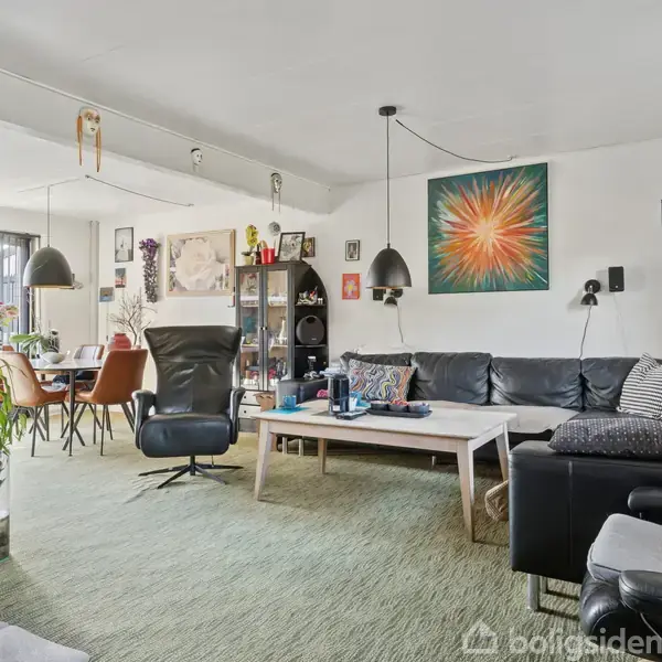 Stue med sort lædersofa og sofabord, dekoreret med kunstværker og planter. En sort lænestol og spisebord til seks personer står i hjørnet. Væggene er lyse.