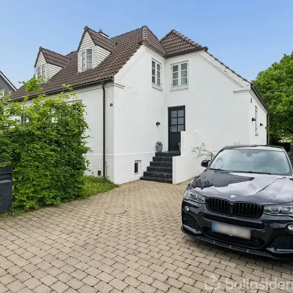 Sort bil parkeret foran hvid villa med brune tegltag. Huset har en trappe ved hoveddøren, og omgives af grønne hække. En anden bygning skimtes i baggrunden.