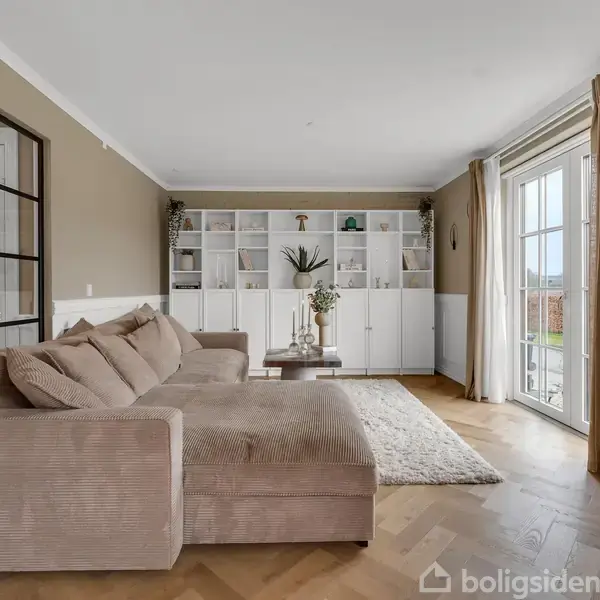 Brun sofa står i en lys stue med parketgulv. Reol med planter og dekorationer fylder væggen. Store vinduer med gardiner slipper lys ind. Fjernsyn hænger på væggen.