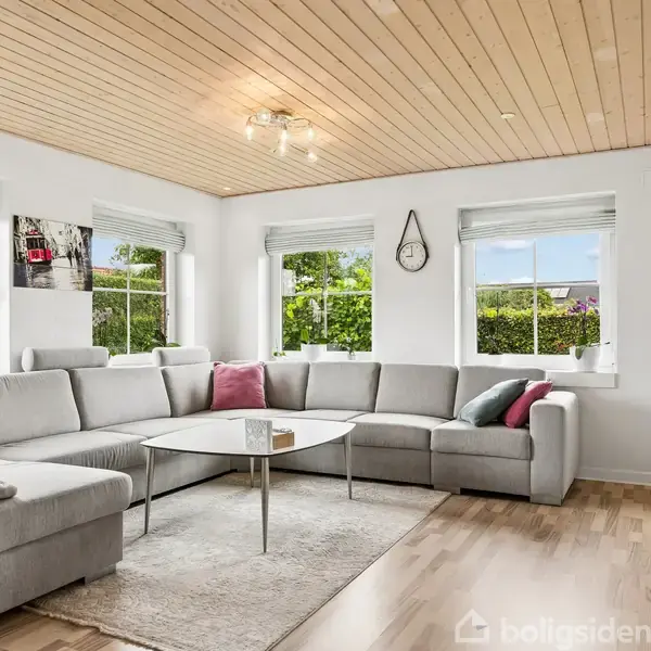 Stor grå hjørnesofa står i lys stue med træloft og vinduer med udsigt til have. En hvid reol med TV og en trekantet vægur hænger på væggen.