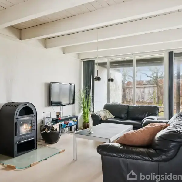 En stue med sorte lædersofaer omkring et sofabord, en vægmonteret fjernsyn, og en brændeovn i hjørnet. Store vinduer og en glasdør giver udsigt til haven.