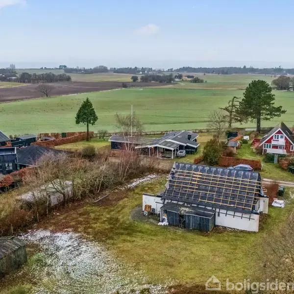 Flere huse ligger spredt i en åben, grøn landbrugsområde med marker i baggrunden. Træer og buske omkranser bygningerne, som har forskellige tagtyper og strukturer.