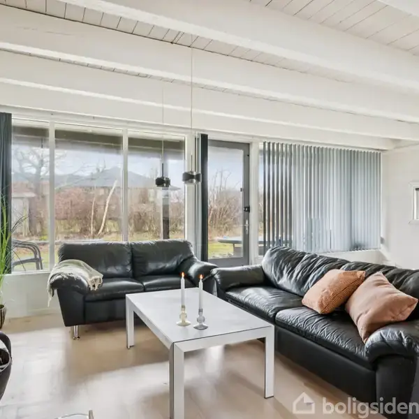 Sort lædermøblement står i opholdsstue. Sofaer omkranser hvidt sofabord med stearinlys. Store vinduer bagved slipper lys ind og afslører udsigt til have med beplantning.