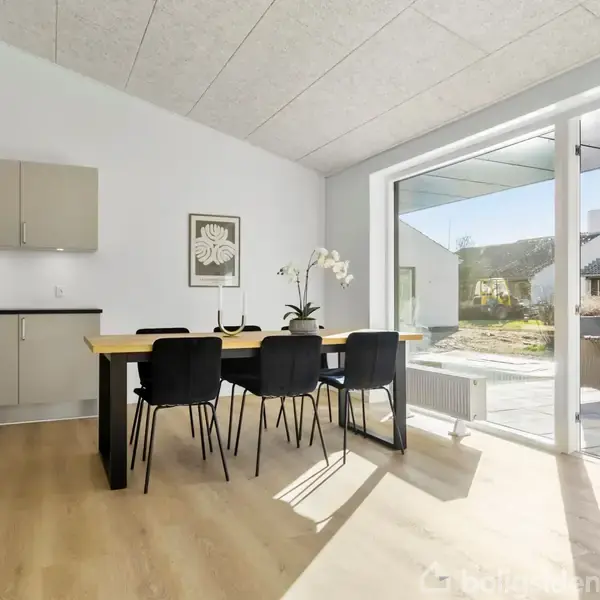 Spisebord med sorte stole står i et lyst køkkenalrum med trægulv. Sollys strømmer ind gennem store vinduer, der åbner ud til en terrasse og have.