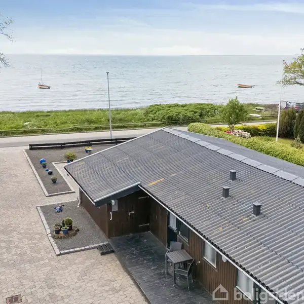 Hus med skifertag ligger ved havet, omgivet af have med buske og træer. Foran huset er der en vej og en stor stenbelagt gårdsplads med blomsterkrukker.