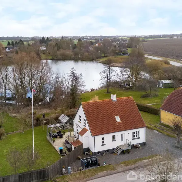 Hus med rødt tag og hvid facade står foran en sø omgivet af træer. Haven har flagstang og terrasse. Landsby set i baggrunden med marker og veje rundt om.