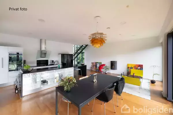 Køkkenalrum med sort spisebord og stole, placeret på trægulv. Køkkenet i baggrunden har hvide skabe og køkkenudstyr. Kunstmalerier og stor gul lampe skaber en moderne atmosfære. Text: "Privat foto".