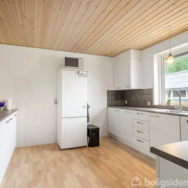 Et moderne køkken med hvide skabe, et køleskab og en opvaskemaskine. Der er en træloft og et stort vindue giver udsigt til en gårdsplads. Lampen hænger over en køkkenbordsplade.