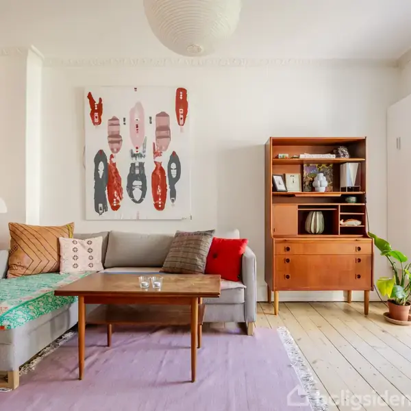 Gråt sofaarrangement med farverige puder står på trægulvet. På væggen hænger en abstrakt kunstplakat. Træskænken ved siden af rummer bøger og planter. Rummet er lyst og indbydende.