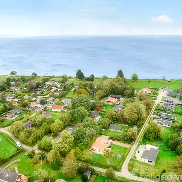 Landsbyområde med huse omgivet af grønne træer nær en kystlinje. Havet strækker sig ud i horisonten under en klar blå himmel.