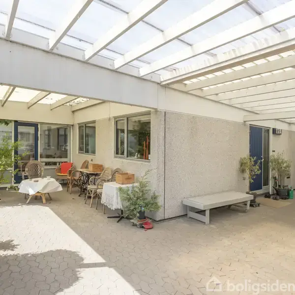 Terrasse med borde og stole, overdækket af et gennemsigtigt tag. Omgivet af planter og en husmur med vinduer. En blå dør fører til indgangen.