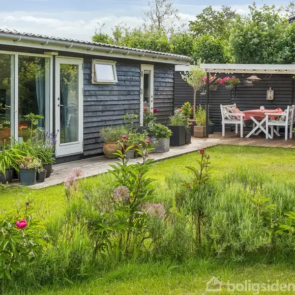 Et sort træhus med glidende døre står i en frodig have. Planter og blomster omkranser en terrasse, hvor et bord og stole er placeret under halvtag.