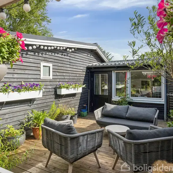 Terrassen har grå lounge-møbler og er omgivet af sorte trævægge. Farverige blomster er plantet i potter og plantagekasser. Sollys skinner gennem træerne fra en blå himmel.