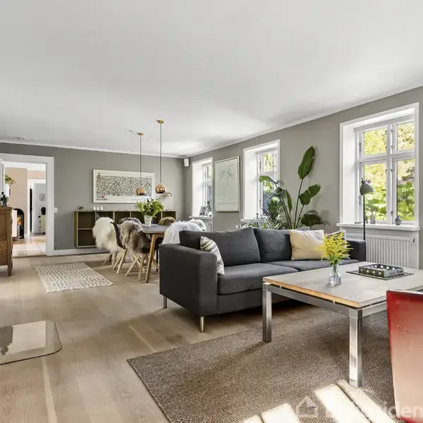 Stue med grå sofa og sofabord; pejs til venstre i hjørnet. Spisebord i baggrunden omgivet af stole; rummelige vinduer og grønne planter giver lys og liv.