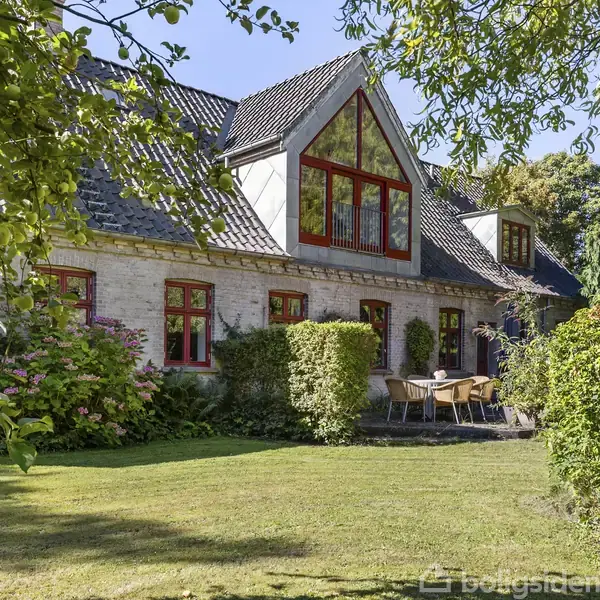 Et charmerende hus med røde vinduesrammer står i en frodig have. På terrassen foran huset står et bord og stole, omgivet af grønne buske under en klar blå himmel.