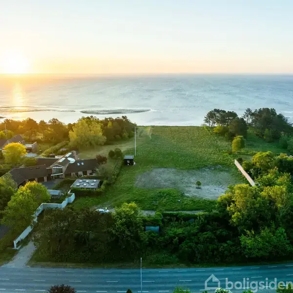 En åben grøn mark ligger ved siden af en række huse nær kysten, mens solen går ned over havet. Træer omkranser området, der skaber en fredelig atmosfære.