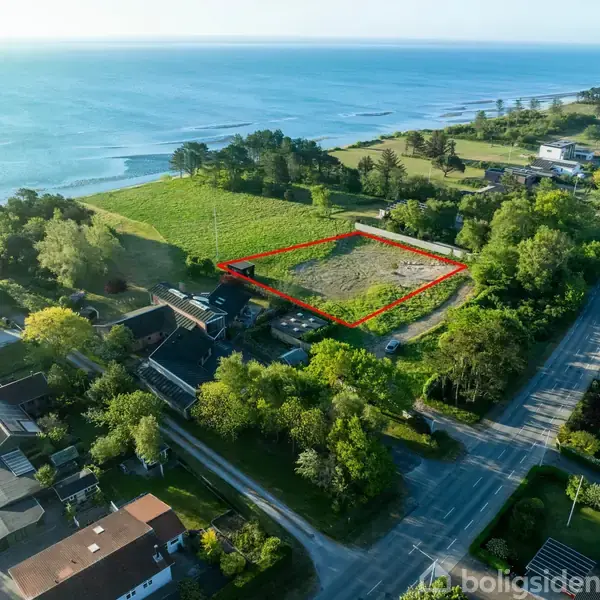 En græsparcel er markeret med rødt i et beboelsesområde ved kysten, omgivet af træer og med udsigt til havet i baggrunden.