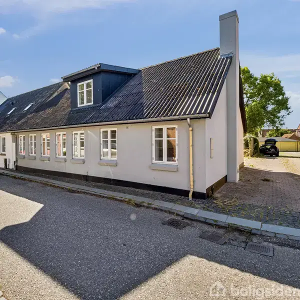 Et langt, hvidt hus med sorte tagsten er placeret langs en stille, brostensbelagt vej. Huset har flere vinduer og en høj skorsten. Solbeskinnet omgivelser med træer i baggrunden.