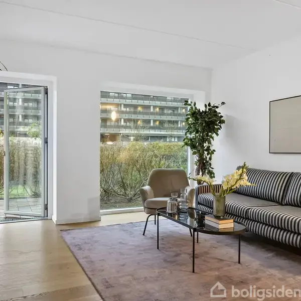 En stue med en stribet sofa ved en glasdør, et sofabord med blomster og bøger, og en lænestol. Udenfor ses en have og en bygning i baggrunden.