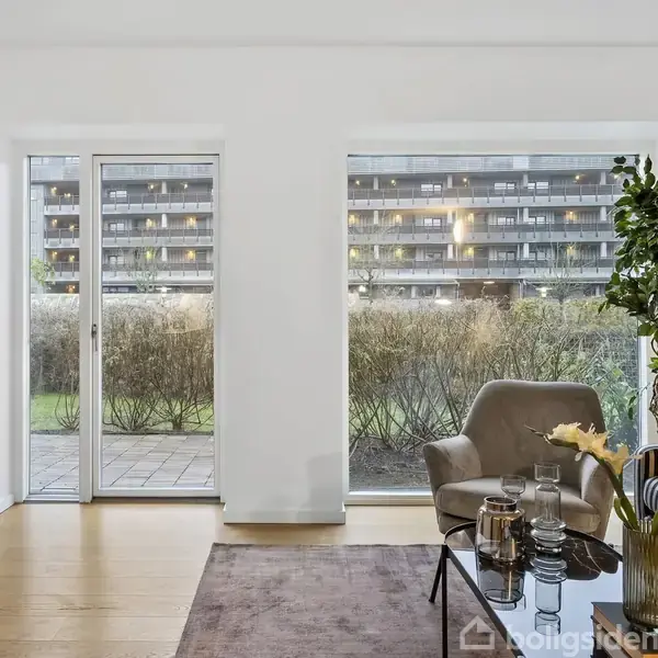 Stue med moderne møbler, herunder en stol og et sofabord med vaser og dekorative genstande. Store vinduer og glasdør fører til en have. Udenfor er der en boligbygning.