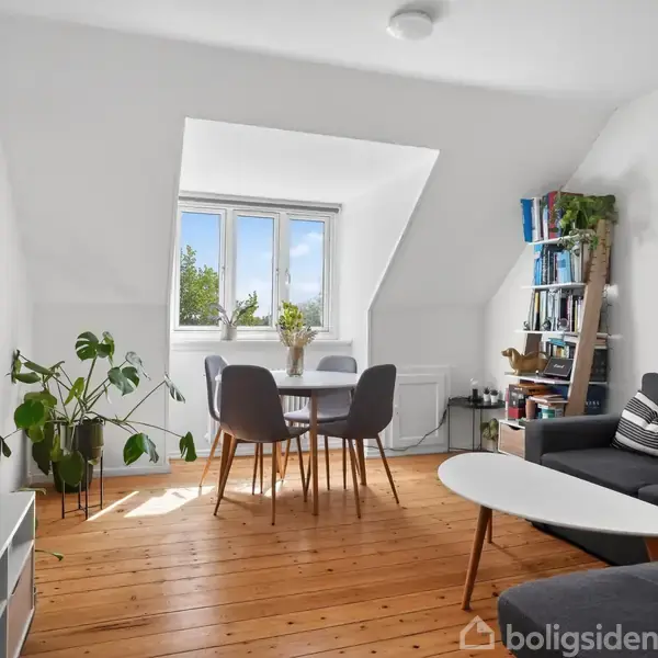 Stue med grå sofa, rundt bord med stole, planter og bogreol. Store vinduer slipper dagslys ind. Boligen er lys og indbydende med trægulv og neutral indretning.