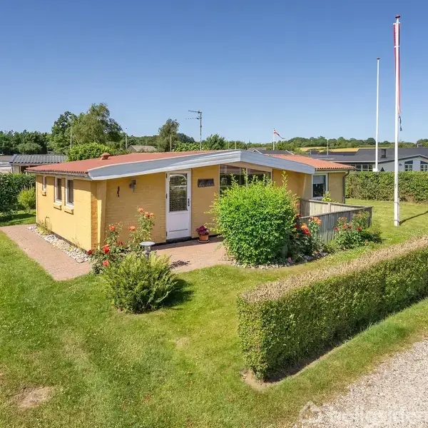 Gul bungalow omkranset af velplejede buske og græsplæne, står under en klar blå himmel. En flagstang til højre og åbne, grønne marker i baggrunden.