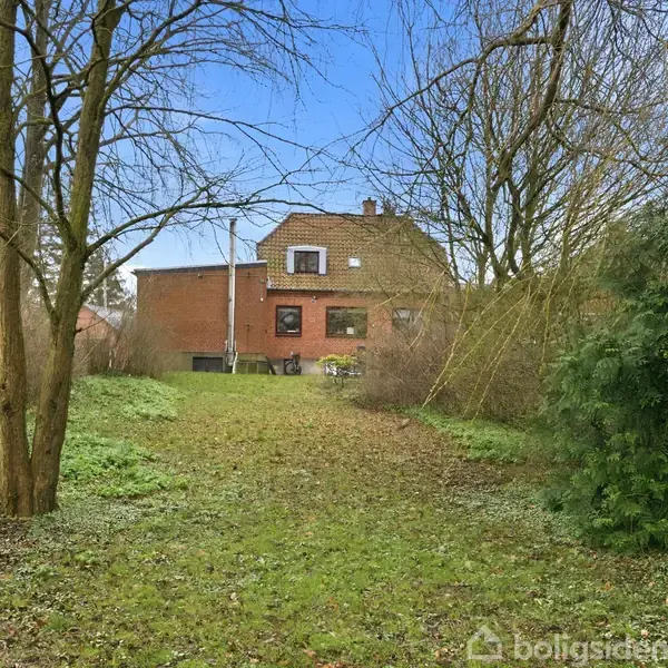 Rødt murstenshus i baggrunden, omgivet af bare træer og grønne buske, på en stor, græsdækket grund under en klar blå himmel.
