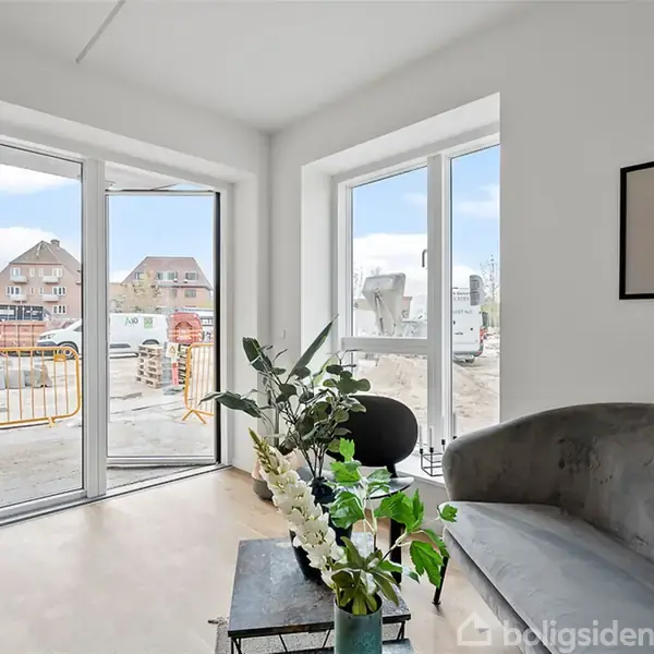 Stue med grå sofa ved glasdøre, der åbner til byggeplads med huse i baggrunden. Plante ved siden af sofabord med grøn vase. Kunstbillede hænger på væggen.