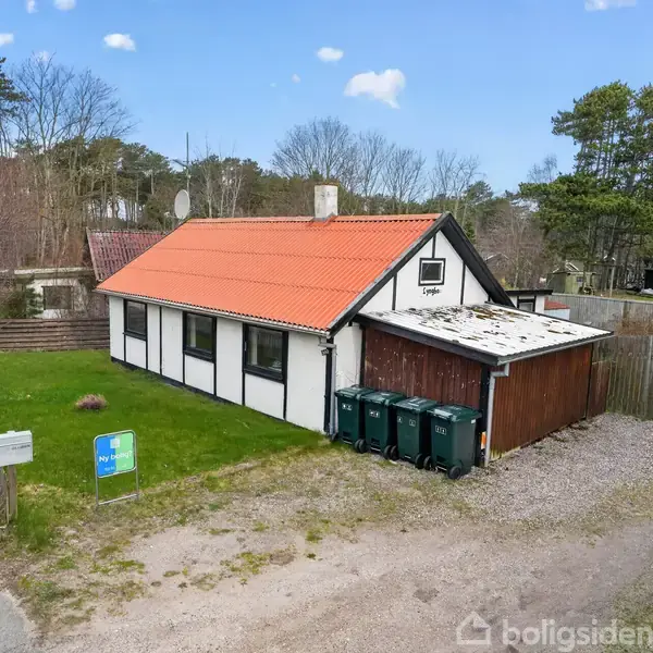 Et lille bindingsværkshus med rødt tegltag står ved en vej. Ved siden af huset er der affaldscontainere. Græsplæne og skovklædt område i baggrunden. To skilte foran huset.