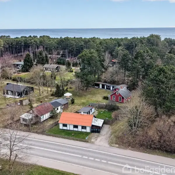 Flere små huse ligger spredt i en skovrig område tæt ved en vej. Havet kan ses i baggrunden, og omgivelserne er frodige og naturlige.