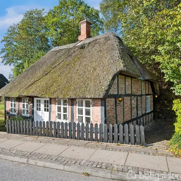 Stråtækt bindingsværkshus står stille ved siden af et fortov omgivet af grønne træer og buske. Huset har et traditionelt, hyggeligt udseende med små vinduer og en lav, sort træstakit.