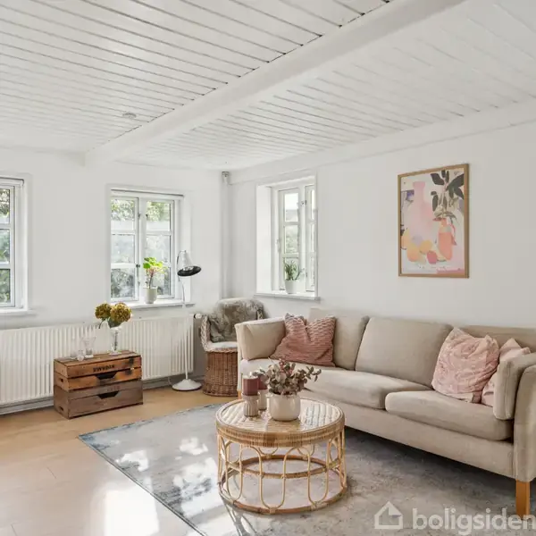 En beige sofa med lyserøde puder står i en lys stue. Et kaffebord med en vase er foran sofaen. Rummet har tre vinduer, en vægplakat og træloft.