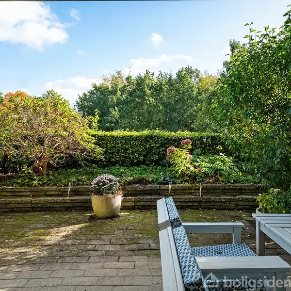En terrasse med et bord og stole står stille omgivet af velplejede buske og potteplanter, set mod en grøn baggrund af frodig vegetation under en blå himmel med skyer.