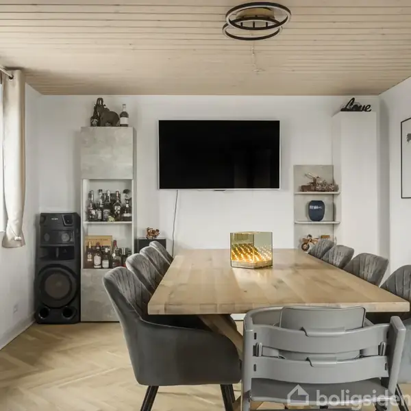 Træbord omgivet af grå stole står midt i et moderne spisestuehjørne. På væggen hænger et tv, mens en reol med spiritus, en højttaler og kortdekorationer supplerer rummet.