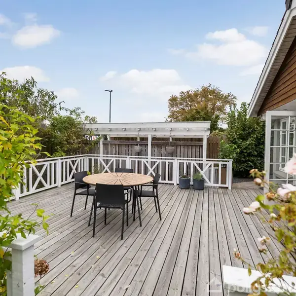 Et rundt bord med fire sorte stole står på en stor træterrasse omgivet af hvidt rækværk. Terrasse ligger foran et hus med store vinduespartier, omgivet af grønne planter.