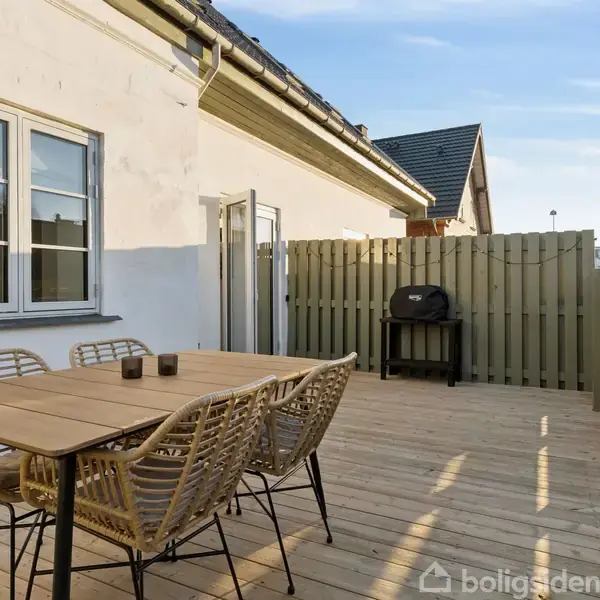 Et træbord med flettede stole står på en træterrasse. En grill står i baggrunden ved et hegn. Vinduer og en dør til huset er til venstre.