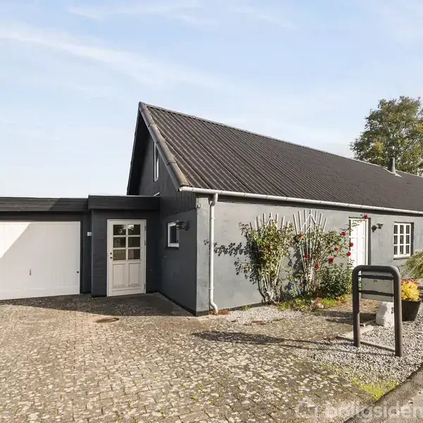 Gråt hus med sadeltag og tilhørende garage. Sollys oplyser gårdspladsen omgivet af grus og beplantning. Skyfri blå himmel i baggrunden.